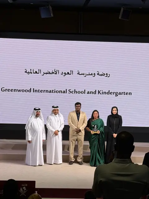 Greenwood receiving the award on stage, the school’s name on the screen behind